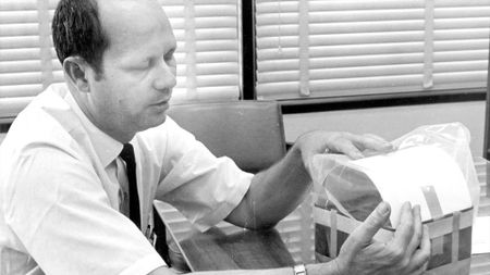 a black and white photo of a man in a white button down shirt and black tie holding a piece of space equipment