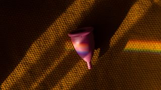 A pink silicon menstrual cup sits on a rubbery brown surface with a rainbow prism of light moving across it 