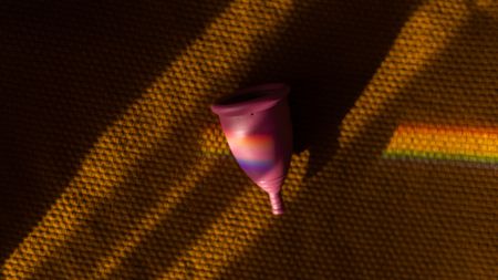 A pink silicon menstrual cup sits on a rubbery brown surface with a rainbow prism of light moving across it 