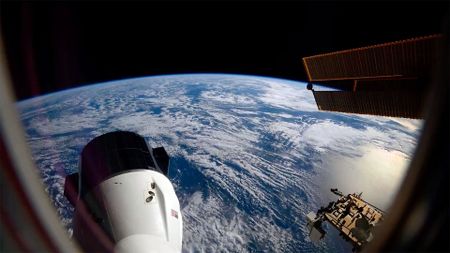 the blue waters and white clouds stretch to out to Earth's horizon, set against the blackness of space; in the foreground, a space capsule extends out from a space station