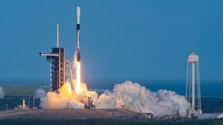 a white and black rocket launches into a clear blue sky from its ocean side launch pad