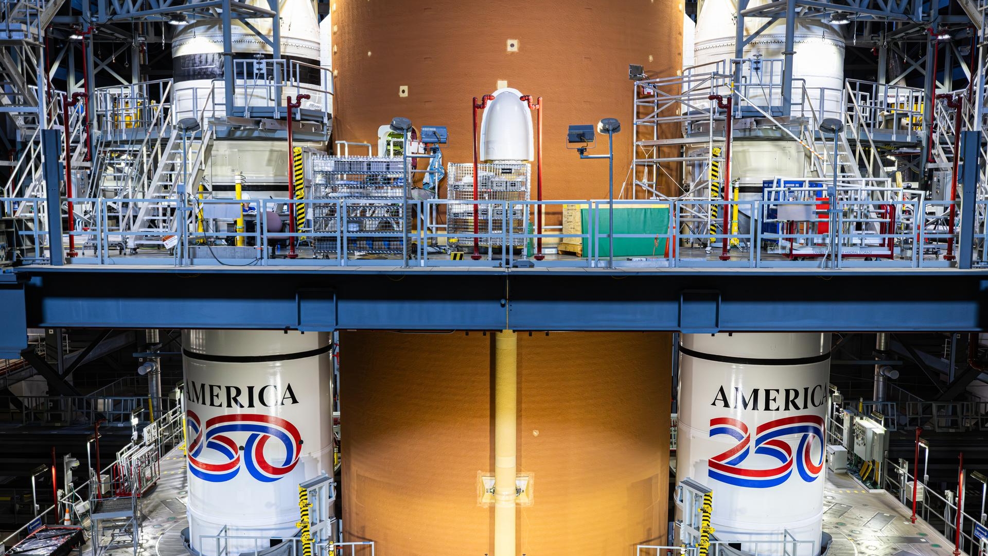 An image from inside a warehouse shows two tall white cylinders on either side of a yellow taller cylinder. The two white cylinders have red, white and blue paint with the words 'America 250' on them.