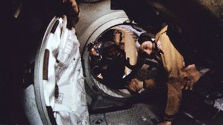 two men floating in space reach out and shake hands across the open threshold of a spacecraft hatchway